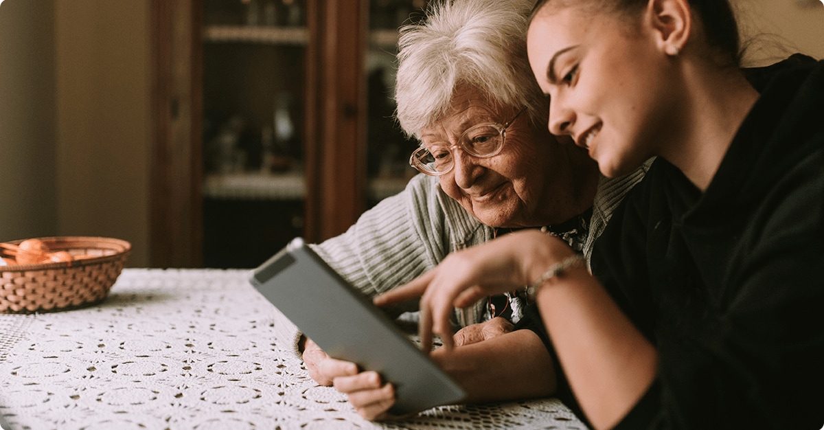 woman sitting at table with elderly relative looking at tablet or smartphone together