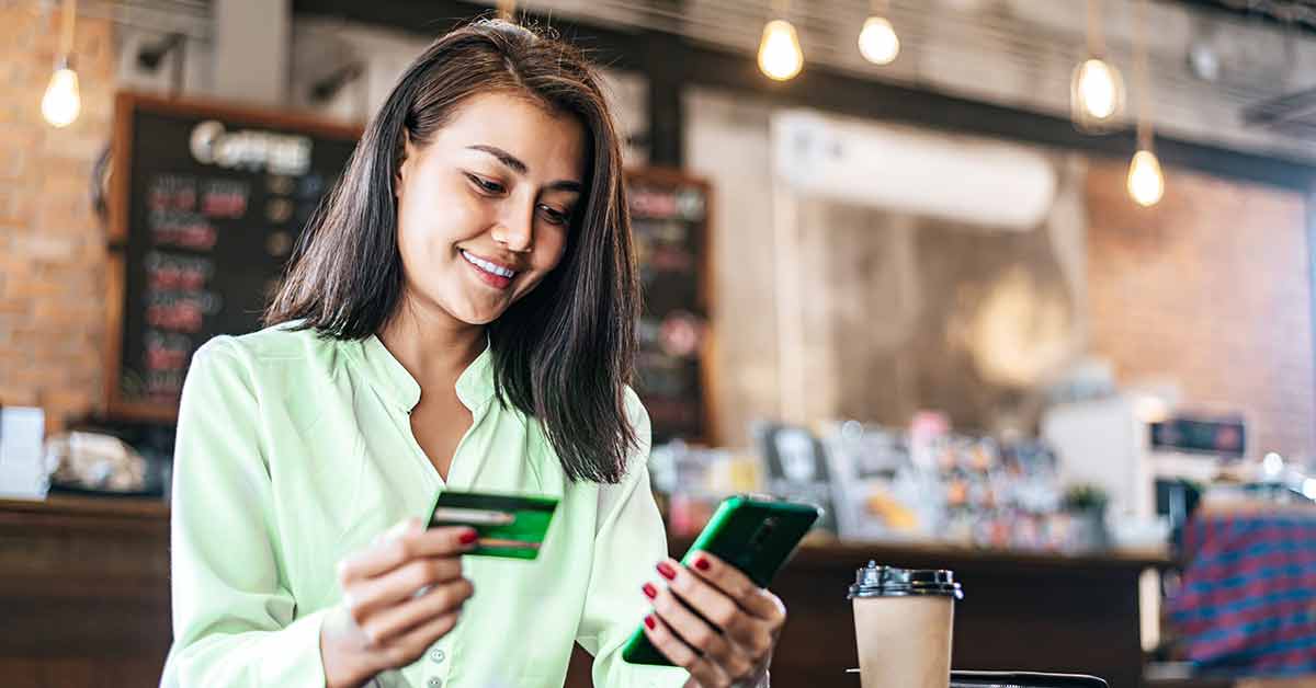 A young woman sits in a coffee shop smiling as she checks her credit card balance using her smartphone.