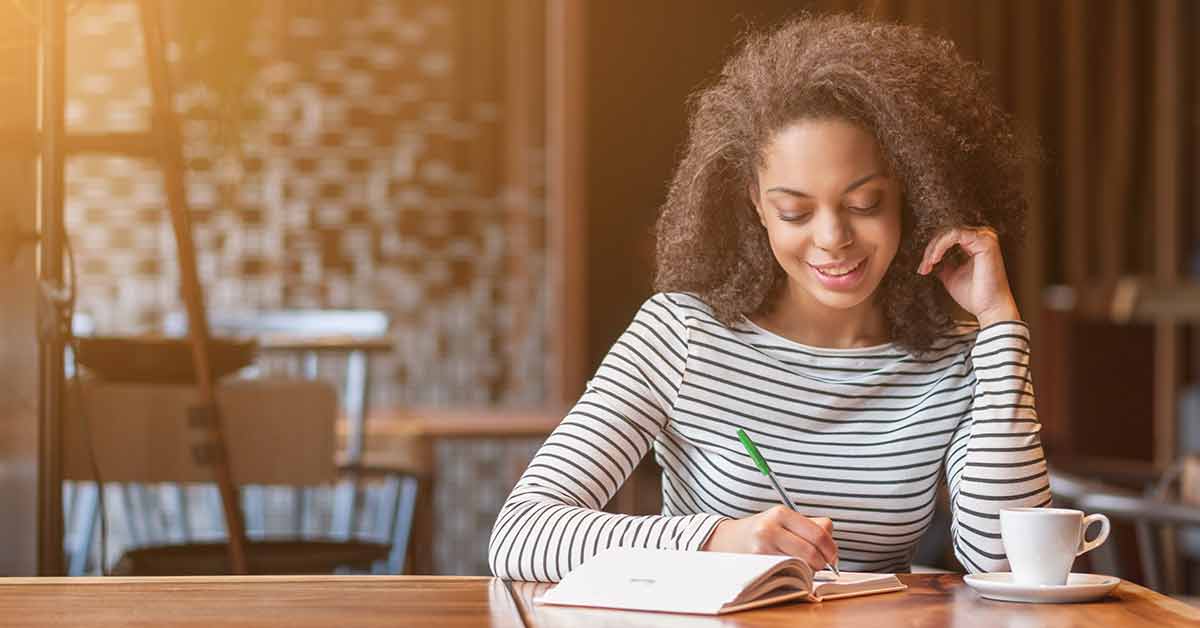 A woman sits in a coffee shop writing her savings goals for the new year in a notebook.